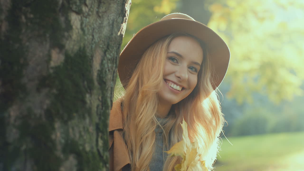 Caucasian young blonde woman wearing hat, holding a yellow leaf and looking at camera behind a tree in the park in autumn
