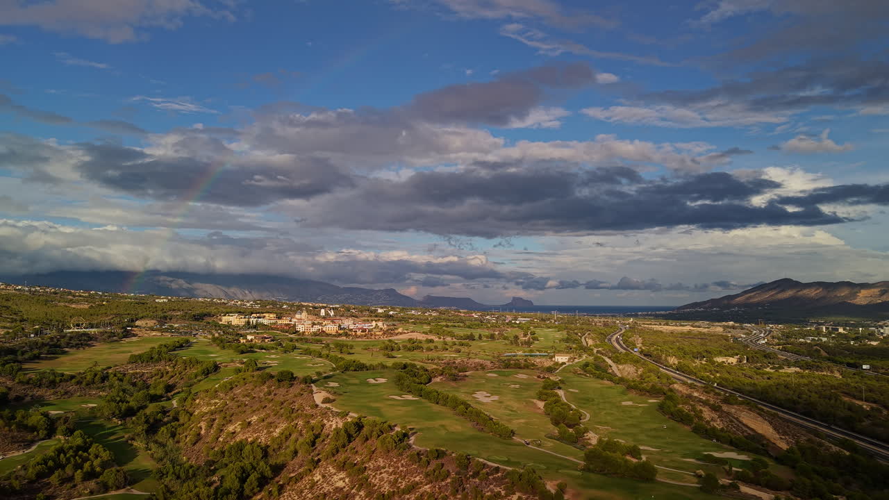 Vista aerea di un paesaggio di campo da golf con campi verdi, colline, autostrada e mare in lontananza sotto un cielo nuvoloso drammatico con arcobaleno
