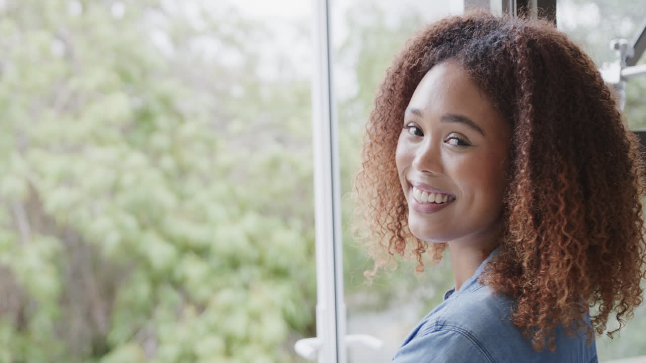 Young African American woman smiles near a window at home, with copy space