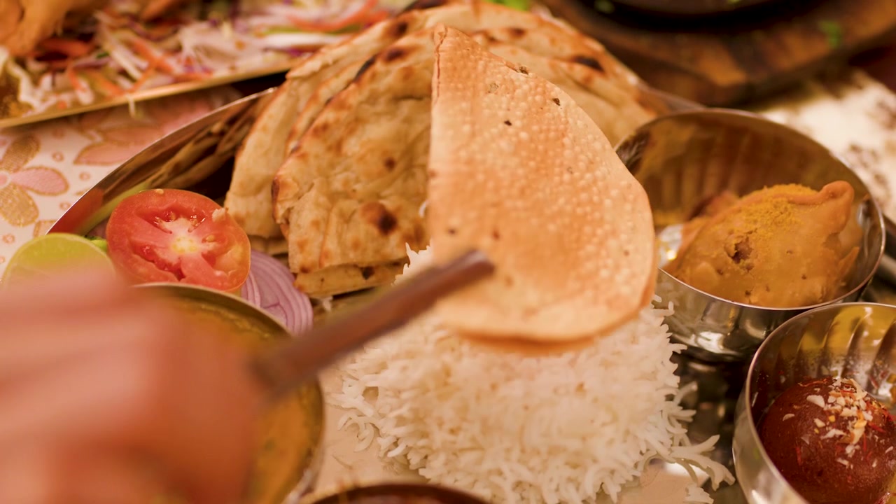 Primo piano di una mano che usa delle pinze per sollevare un papadum da un vario pasto thali indiano.