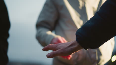 close-up of three individuals stacking their hands together in a gesture of unity and teamwork, with a blurred background that includes someone wearing a gray jacket