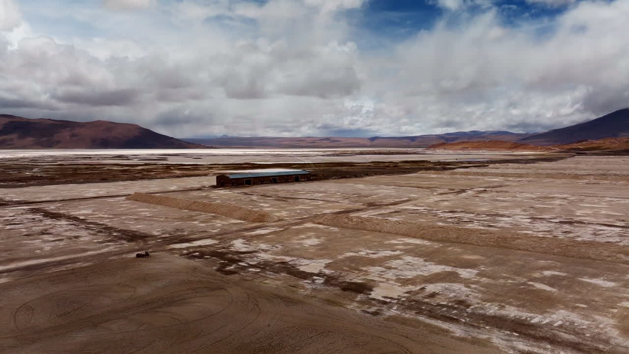 Borax mine at Laguna Capina, evaporation ponds, industrial building, and mountains under cloudy sky, Bolivia. Aerial forward