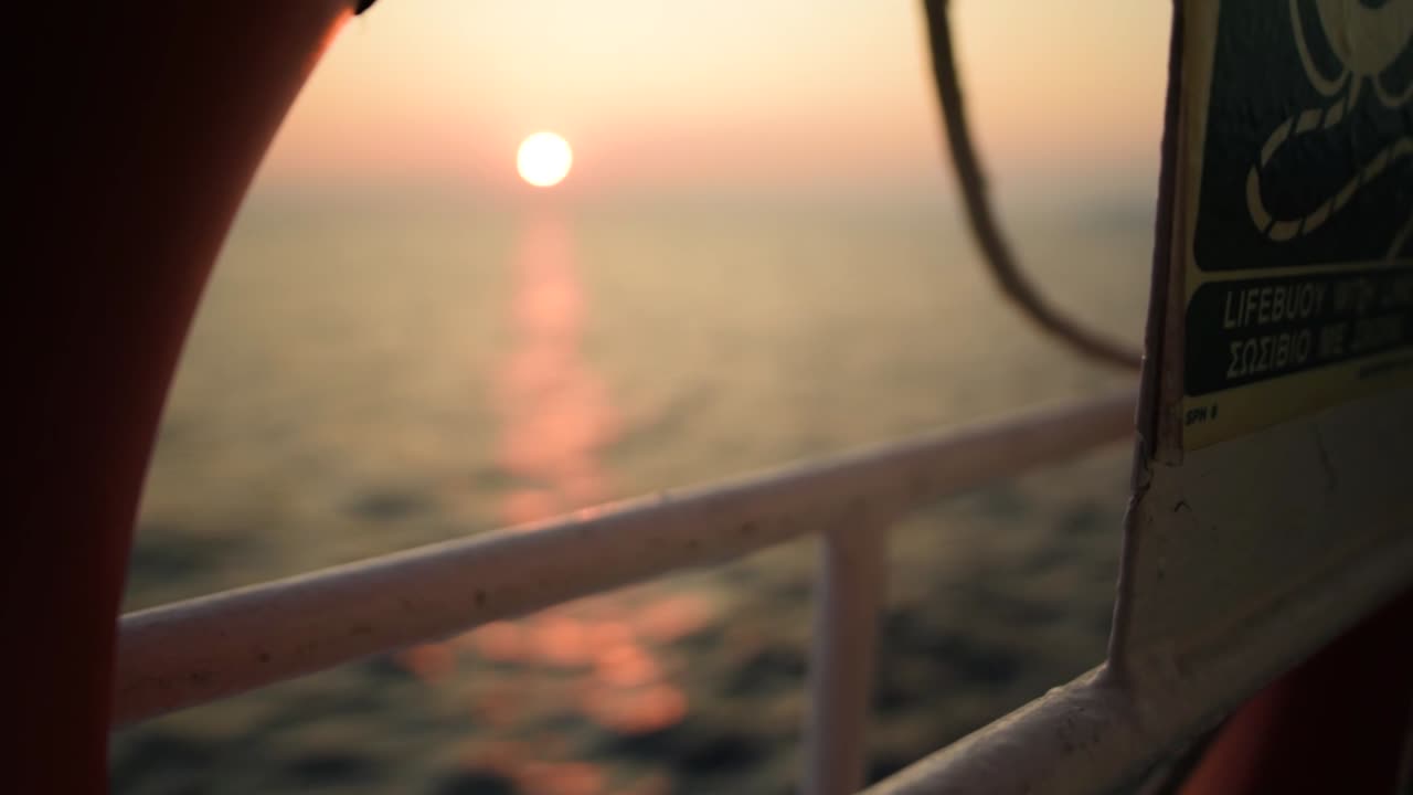 Rack-Fokus-Aufnahme auf ein Schiff, das bei Sonnenuntergang auf dem Meer fährt, mit der Sonne im Hintergrund und einem Rettungsring im Vordergrund, Zeitlupe, Griechenland