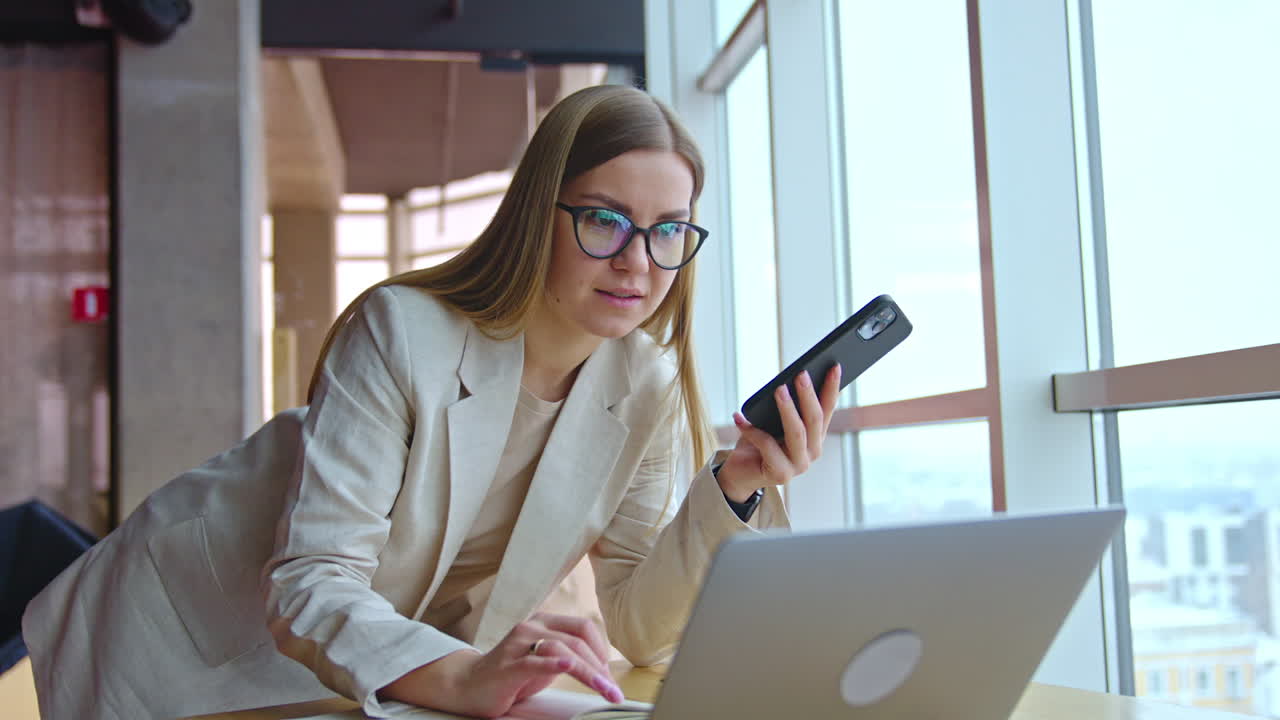 Long-haired woman in glasses uses phone and laptop at once. Lady stands above the desk and types something on computer.