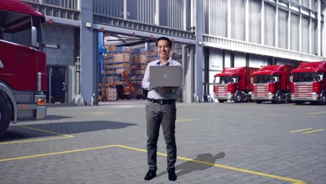 full body of an asian male professional worker standing with his laptop, outside of logistics distributions warehouse, he is looking at the camera with a smile