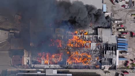 Black-smoke-rise-as-inferno-with-red-hot-flames-ravages-warehouse,-top-aerial