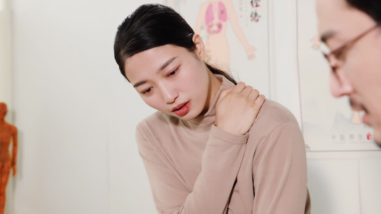 理学療法士の男性医師が、慢性疾患、健康上の不調、症状について話す女性に耳を傾け、患者の肩や背中の痛みの話を聞きながらメモを取り、病歴を記入している