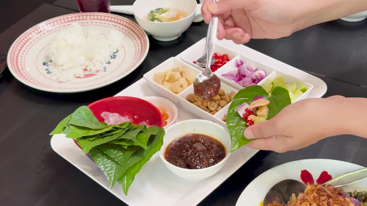 Hands assemble Miang Kham savory leaf wraps using fresh betel leaves, assorted fillings, and sauce on a tray in bright indoor lighting with a static camera