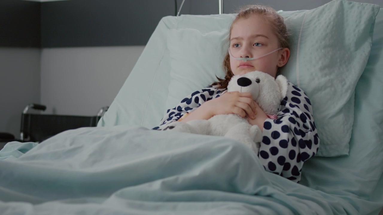 Sad Young Girl With Nasal Cannula Holding Teddy Bear in Hospital Bed