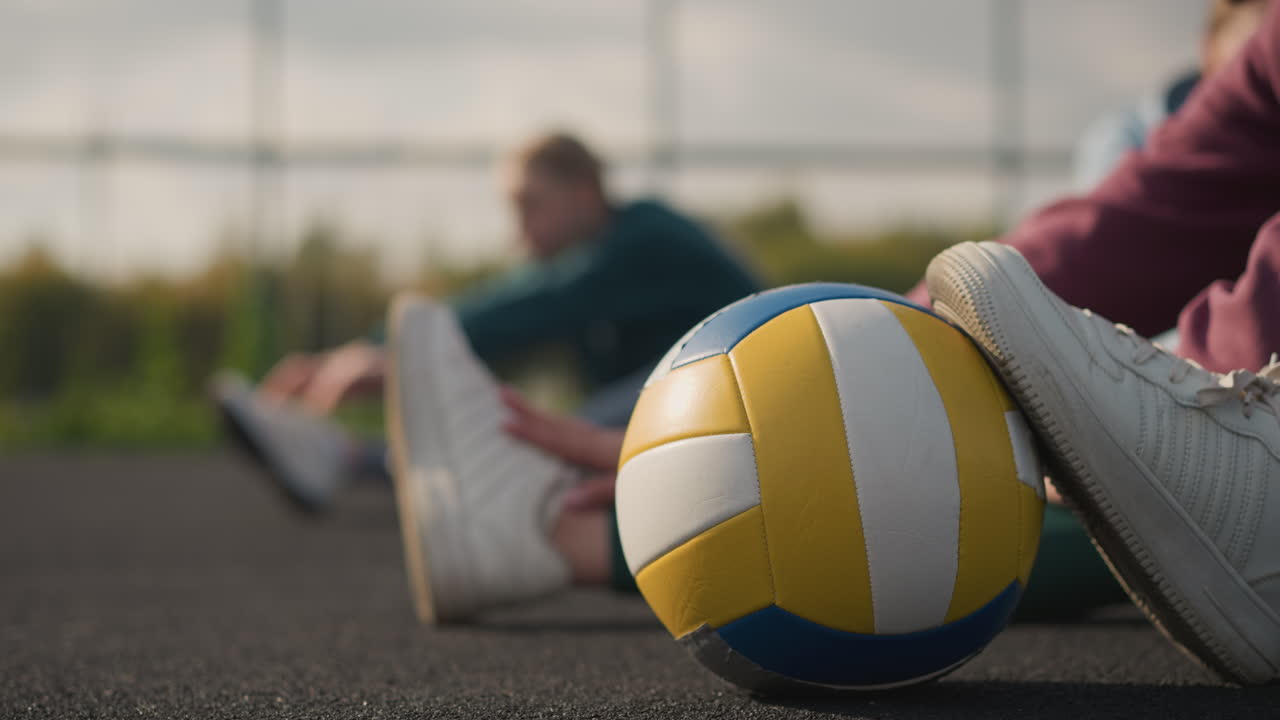 야외 코트에서 누군가의 발 에 있는 배구공의 클로즈업, 배경에 어있는 여성의 흐릿한 시야, 스포츠 훈련 환경에서 운동 세션을 포착합니다.