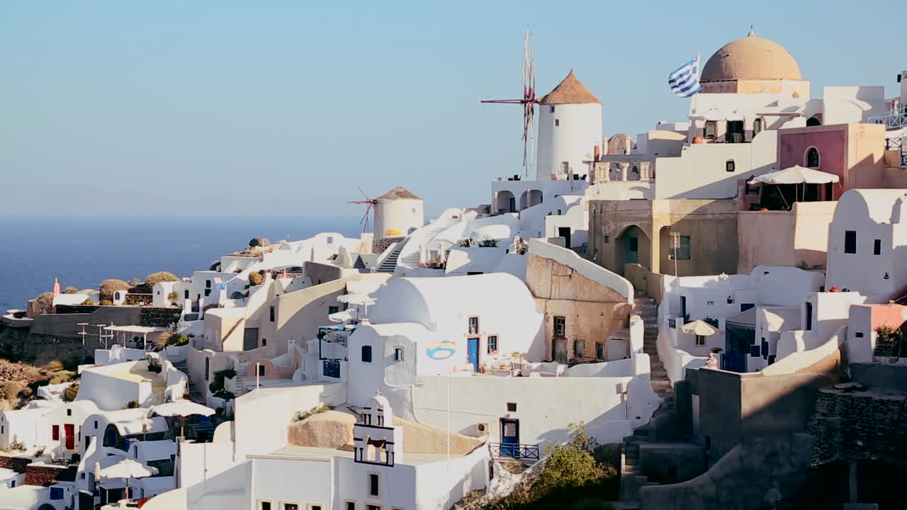 edificios blancos y molinos de viento bordean las laderas de la isla griega de santorini con una bandera griega en la distancia