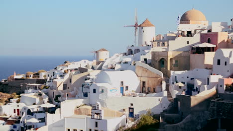 Los-Edificios-Blancos-Y-Los-Molinos-De-Viento-Se-Alinean-En-Las-Laderas-De-La-Isla-Griega-De-Santorini-Con-Una-Bandera-Griega-En-La-Distancia