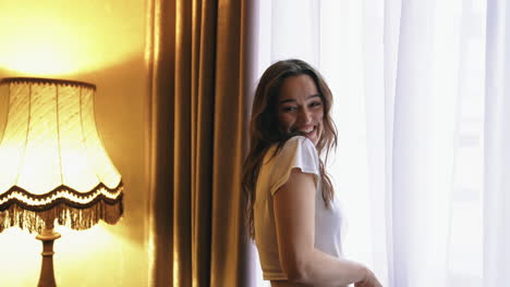 Young happy smiling woman looks out from a window of a hotel room. Large windows in a modern hotel room with up to date interior. Happy holidays.