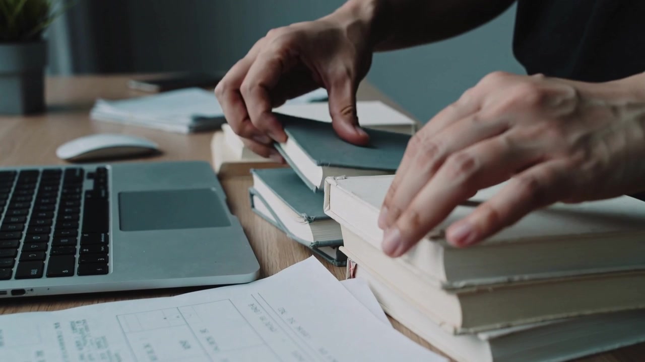 Reaching from right, hands wearing short sleeve dark shirt arranging books by laptop, sorting forms