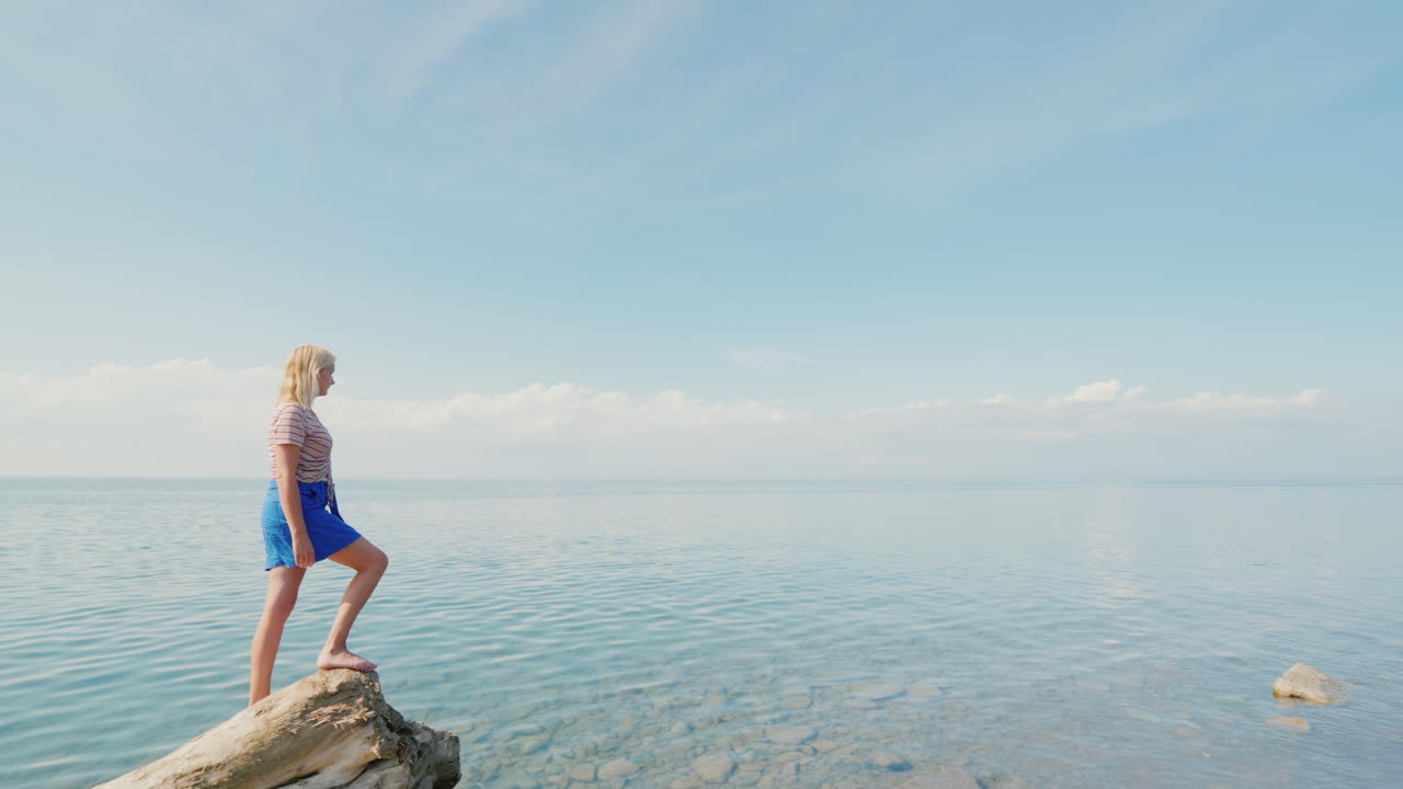 활동적인 여성 여행자는 통나무 가장자리에 서서 잔잔한 바다와 파란색의 아름다운 전망을 감상합니다.