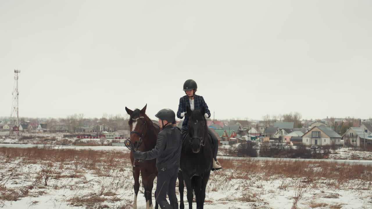 Due cavalieri a cavallo in un campo innevato, una scena di educazione equestre in tandem con un istruttore a terra che guida lo studente. Cielo coperto e un villaggio all'orizzonte, giacche invernali e caschi, briglie e selle, atmosfera calma.