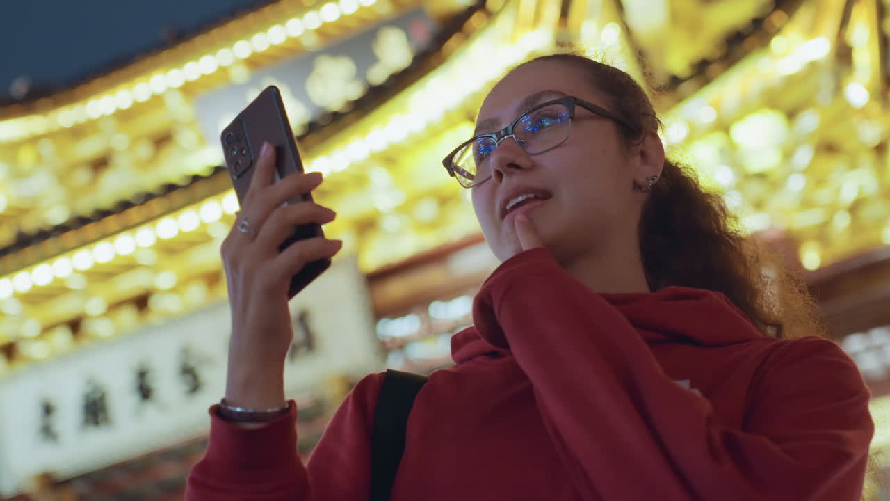 Ung kvinne direktesender ved et gyllent tempel om natten, engasjerer følgere med telefon, rød hettegenser og briller, selvsikkert smil og livlige gester, ornamentert opplyst tak og lanterner, på lokasjon