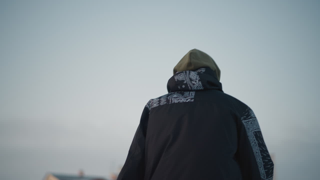rear view of young boy skating under bright winter sky in patterned jacket with blurred residential building backdrop, smooth ice surface reflecting pale light and creating calm seasonal atmosphere