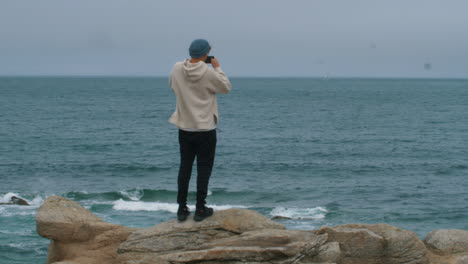 casual looking young man with blue hat taking photo with smartphone in front of ocean