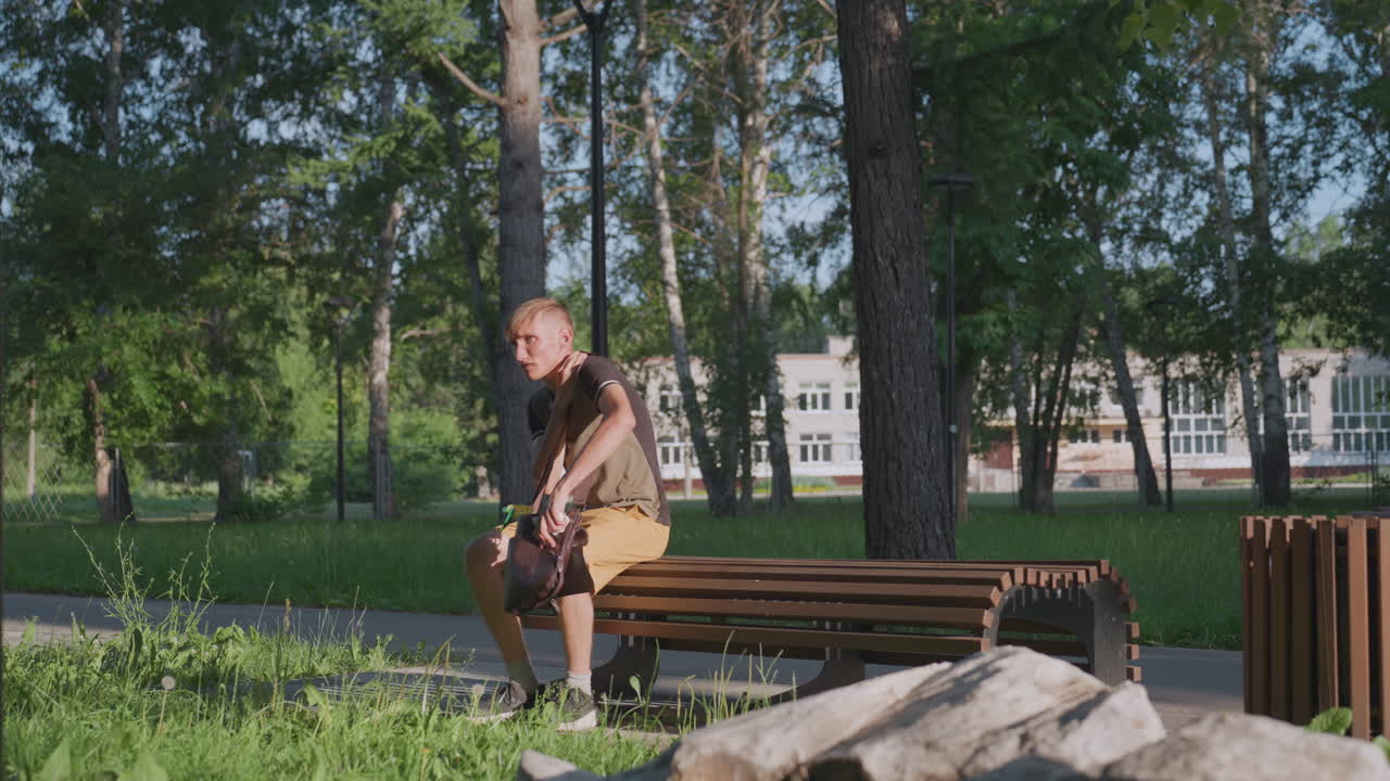 Individual Inspects Plastic Container, Man Carefully Looks At Plastic Bottle Near Bench In Sunlight, Individual Bends To Observe Plastic Bottle Beside Bench With Trees And Buildings Behind