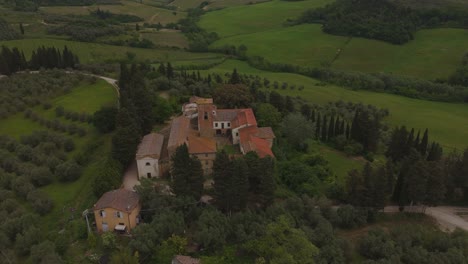 Drone shot orbiting an old Tuscan stone complex with a central bell tower, surrounded by olive trees, rolling green hills, and cypress trees. Overcast light enhances the rustic and peaceful atmosphere