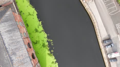 drone footage captures a top-down view of the river aire, adjacent buildings, parked cars, and vibrant green riverbank vegetation in daylight