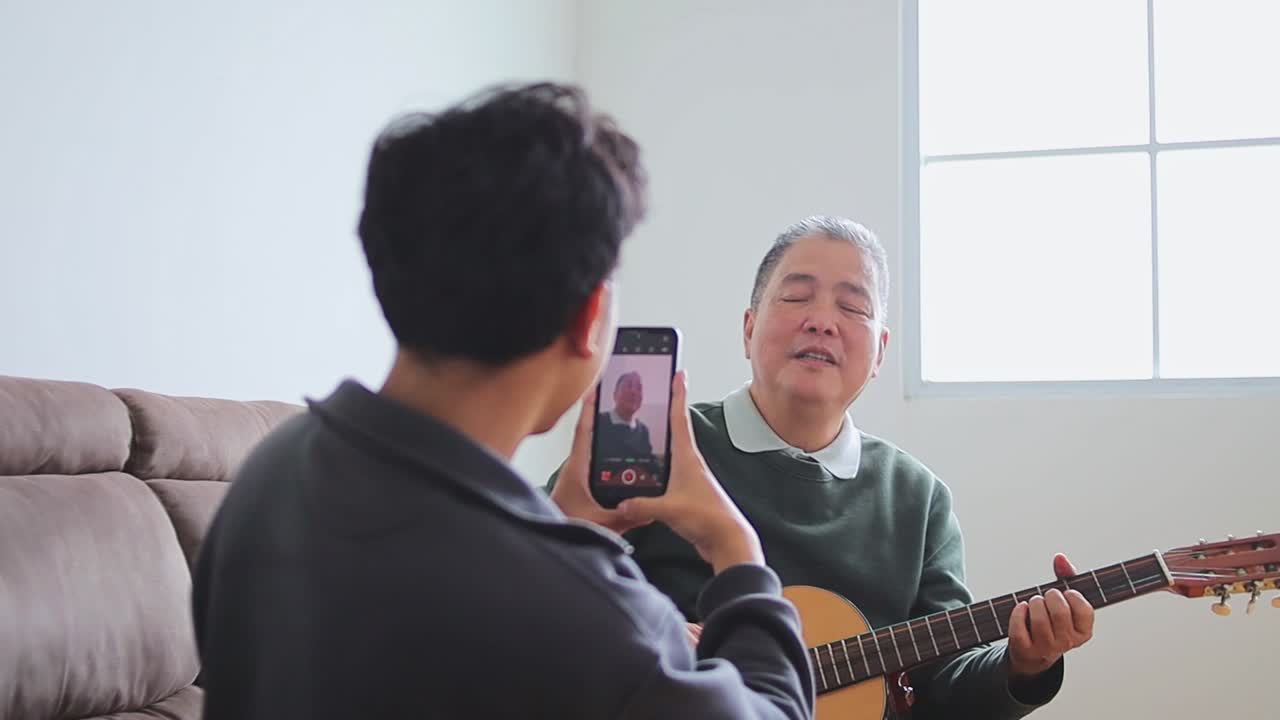 Grandson recording grandpa playing guitar with smiling on comfortable couch