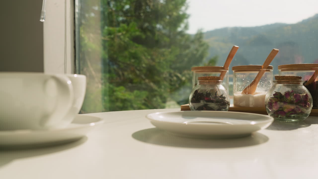 젊은 여성은 테이블에 차를 마시기 위해 접시를 놓습니다. 아내는 테이블 아침 식사를 준비합니다. 약초 차 컵이 이미 위치합니다. 양육하고 활력을 불어넣는 아침 의식.