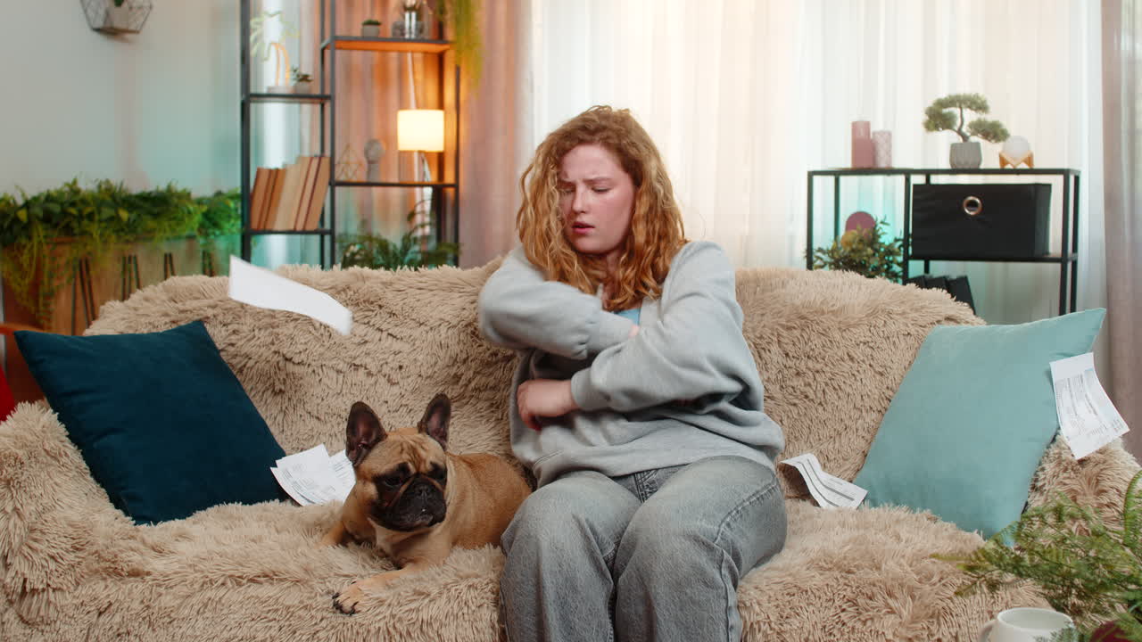 Young woman throws bills on home sofa in frustration while pug dog watches with confused expression