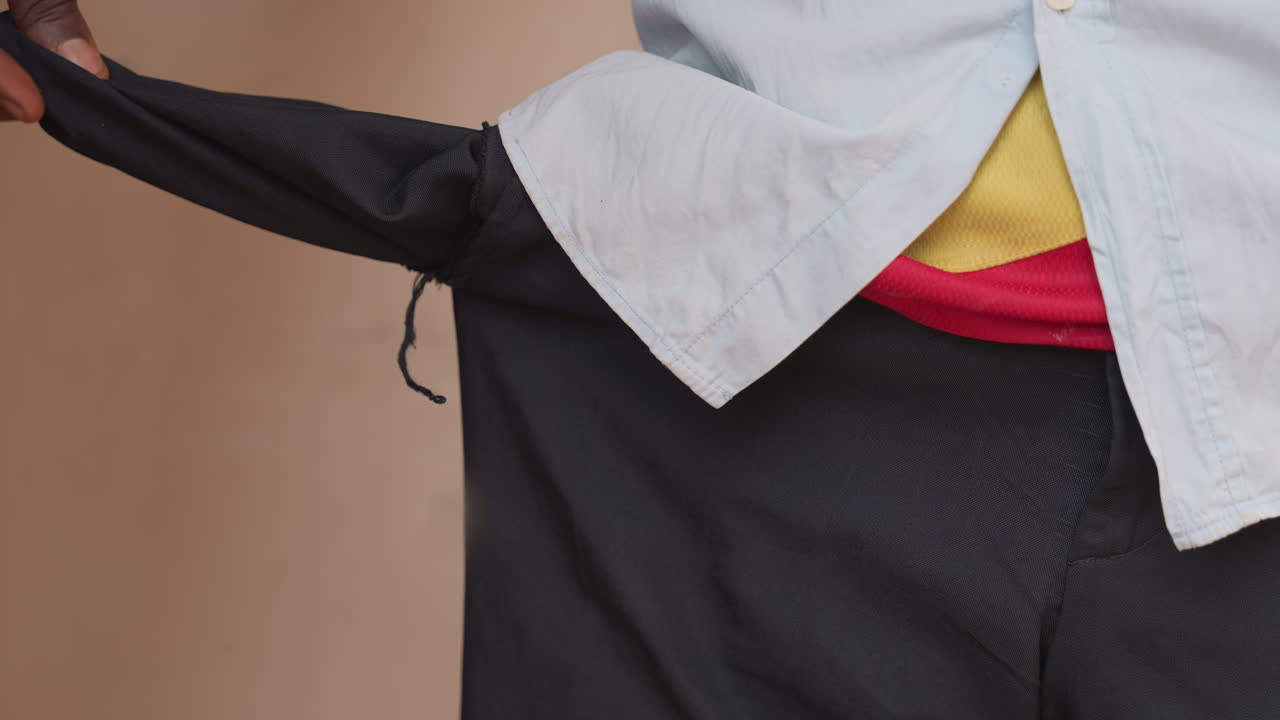 Hombre negro inspeccionando un bolsillo roto, primer plano mostrando camisa blanca y camiseta interior roja, tirando del forro de la cintura para evaluar el daño, ambiente de reparación doméstica, manos revisando la costura, interior modesto, cotidiano