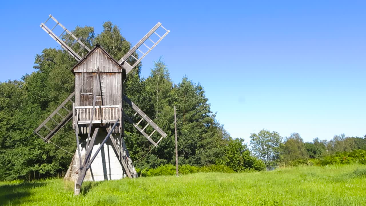 Low angle pan shot of traditional Danish post windmill, built from wooden boards standing in sunlit summer green grass meadow landscape at rural forest edge, clear blue sky. Agricultural tradition