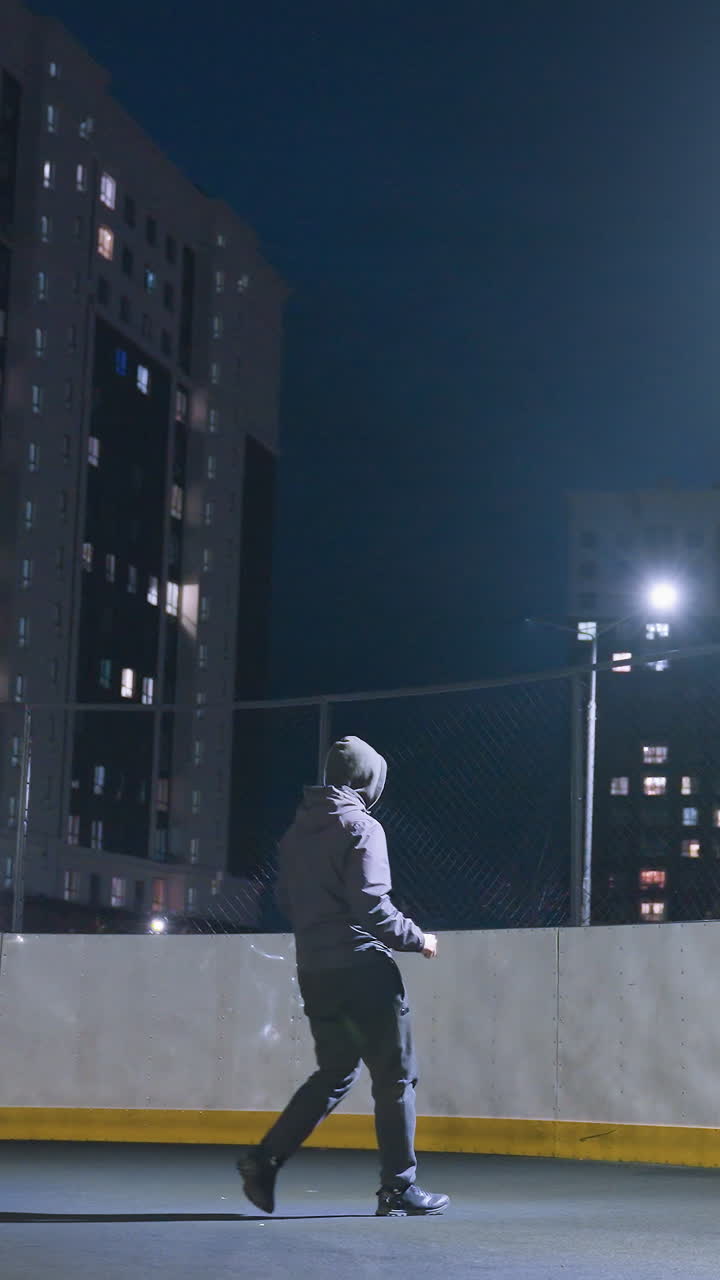 Back view of athlete in hoodie kicking soccer ball against wall under bright stadium lighting at night, ball reflects silhouette on wall with urban cityscape and illuminated apartment buildings