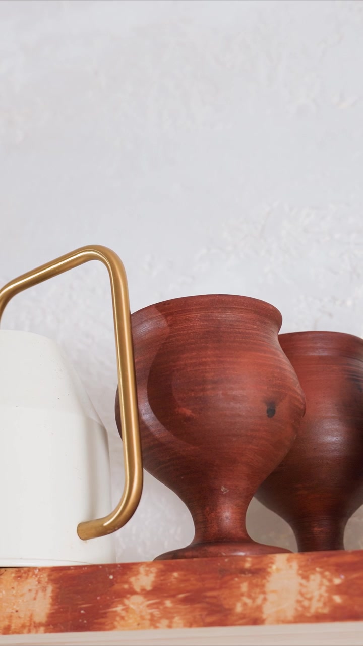 Close up of white ceramic pitcher with golden handle next. Objects are stationary with no motion set. Shelf has rustic reddish brown appearance with visible texture