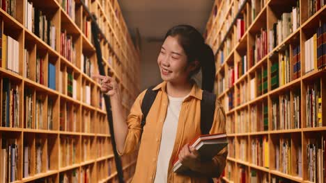 asian woman student with a backpack and some books smiling and pointing to side while standing in the library