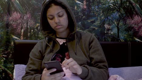 close up shot of a girl in a brown color hoodie sitting in her room and surfing mobile.