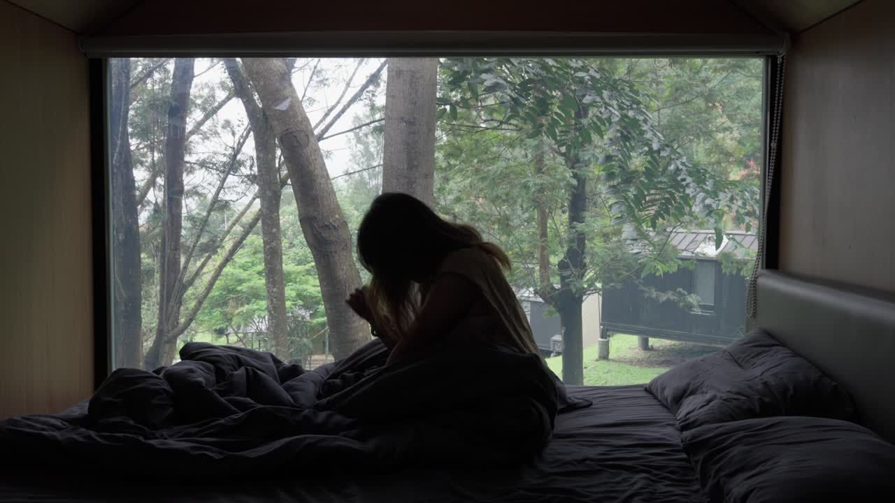 Young woman stretching after waking up in bed, with a nature view outside the window