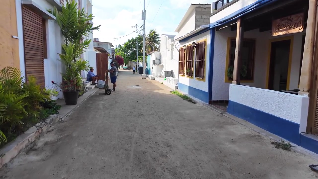 steady tracking shot capturing the contrast between local homes and coastal inns along a sandy street in a vibrant Caribbean village in Venezuela