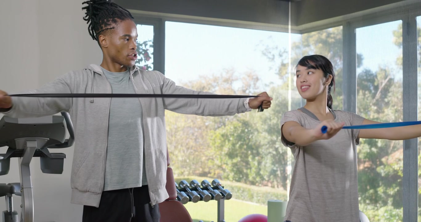 Diverse workout partners wearing sportswear holding bands at chest in gym, repeating pulls for back