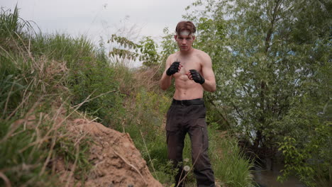 warrior standing shirtless on grassy riverbank preparing to throw punches with gloved fists, rubbing under nose with intense focus, surrounded by wild vegetation under cloudy sky in rugged terrain