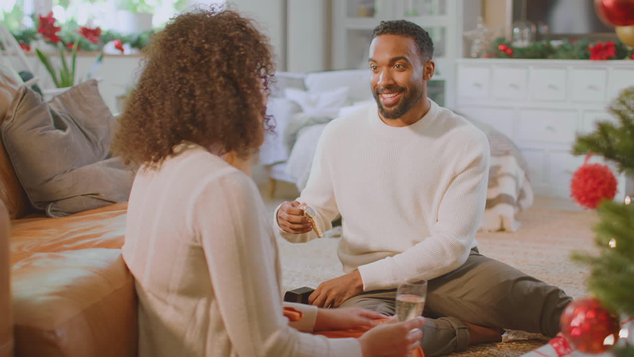 Loving Couple Drinking Champagne As Man Opens Gift Of Wristwatch Around Christmas Tree At Home