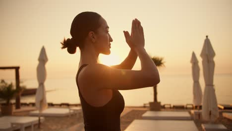 A-brunette-girl-with-her-hair-tied-in-a-bun-in-a-black-top-is-meditating-on-a-Sunny-beach-in-the-morning-and-puts-her-hands-in-the-Namaste-position