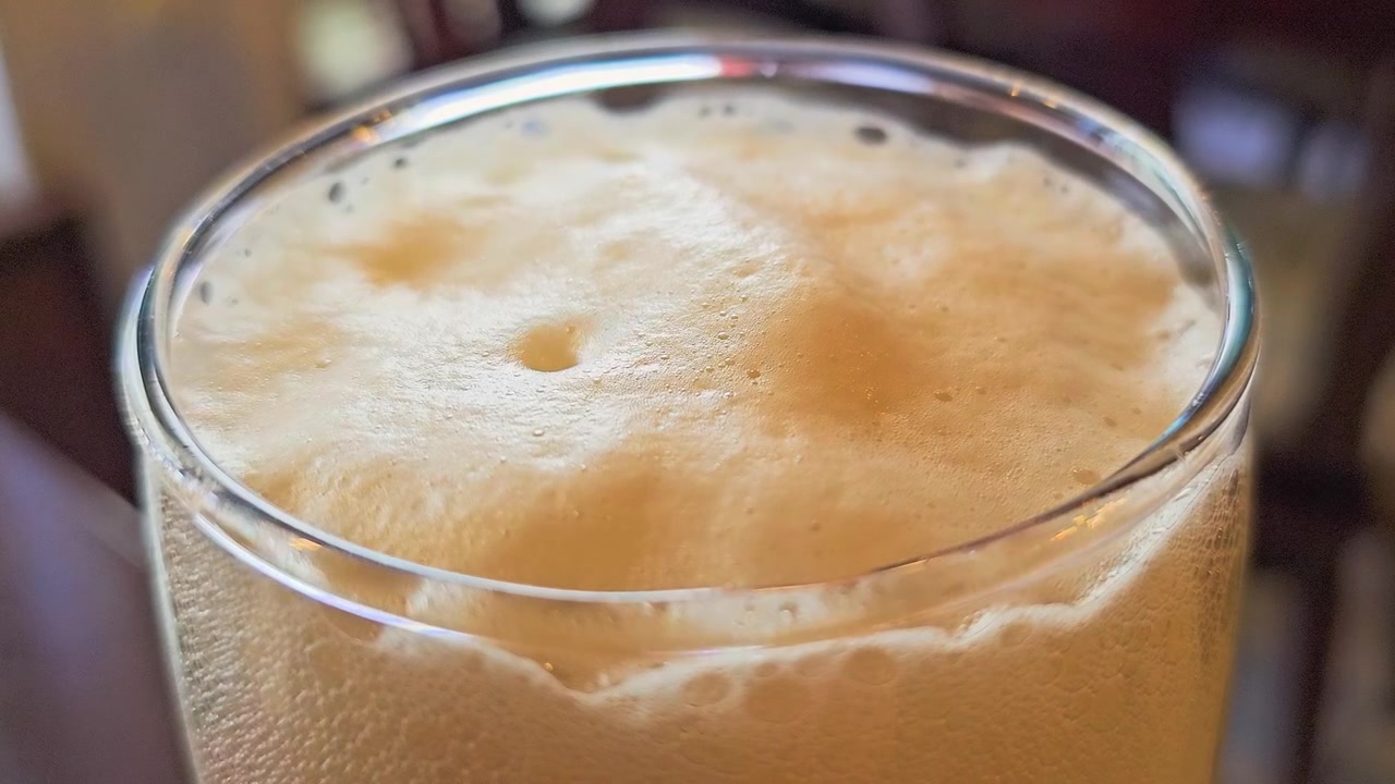 Sıcak bir pub veya restoran ortamında, altın rengi köpük, baloncuklar, buğu ve sığ alan derinliği ile köpüklü bir bira bardağının yakın çekimi, içecek ve gece hayatı stok videosu için mükemmel