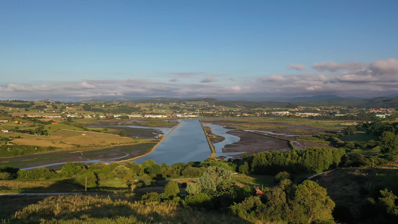 Flight over mouth of a river where you can see a garden area next to the old banks of the estuary surrounded by rural houses and green meadows and mountains on a sunny summer day in Cantabria Spain