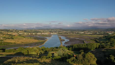 Flight-over-mouth-of-a-river-where-you-can-see-a-garden-area-next-to-the-old-banks-of-the-estuary-surrounded-by-rural-houses-and-green-meadows-and-mountains-on-a-sunny-summer-day-in-Cantabria-Spain