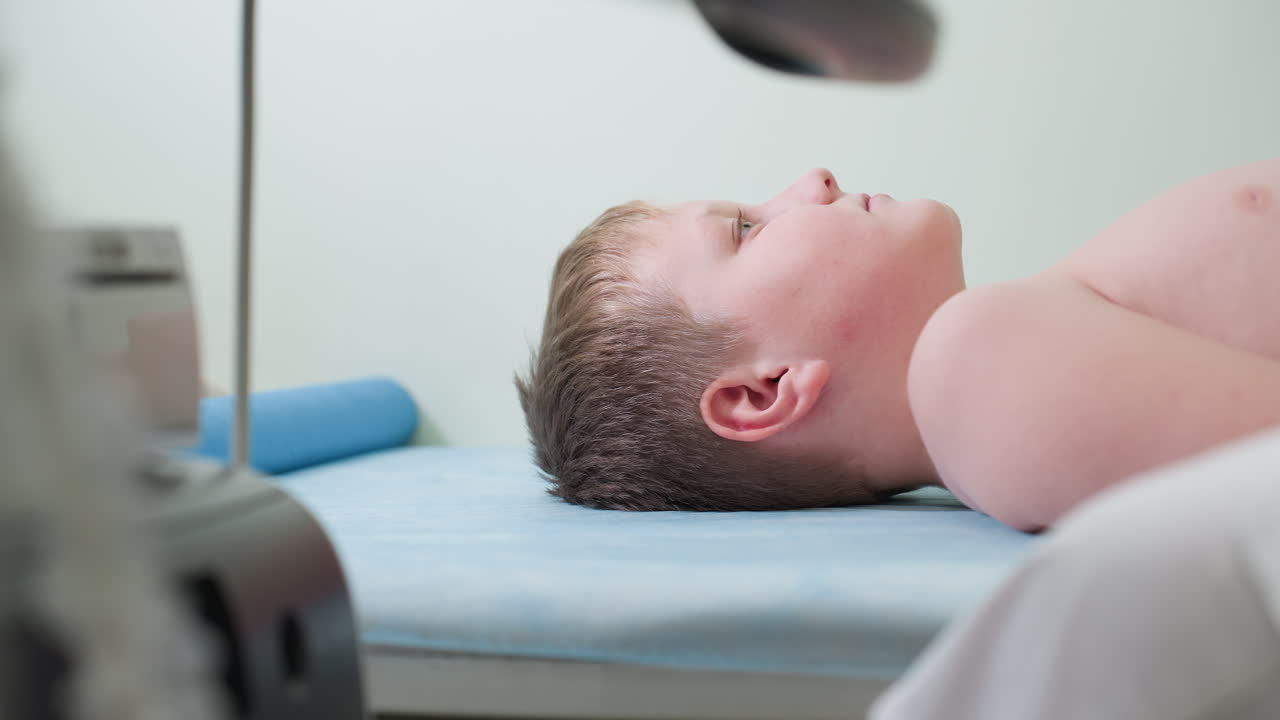 Teenage boy lying straight on clinical bed blinking occasionally during medical examination, appearing calm and relaxed while surrounded by clinical instruments and monitored by healthcare worker