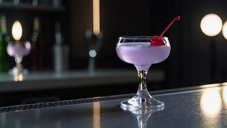 pink aviation cocktail resting on wooden bar counter, reflecting warm ambient lighting with brandied cherry garnish casting soft glow during evening social gathering
