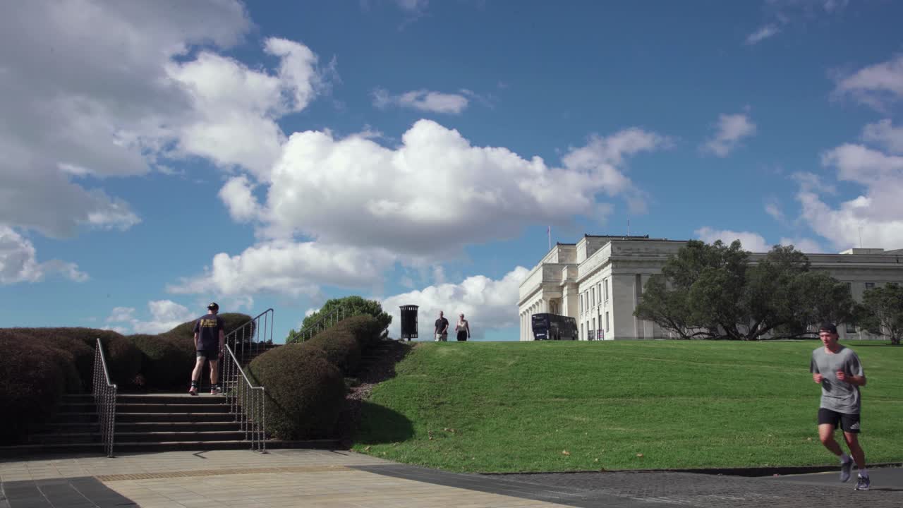 People walk and jog near steps and a grassy hill in front of the Auckland War Memorial Museum on a picturesque day