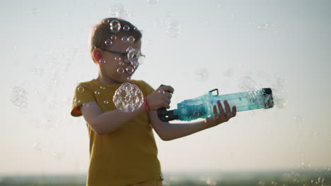 young boy wearing a yellow shirt and shorts, with glasses, stands outdoors, holding a plastic container that releases bubbles into the air. in a bright, open field