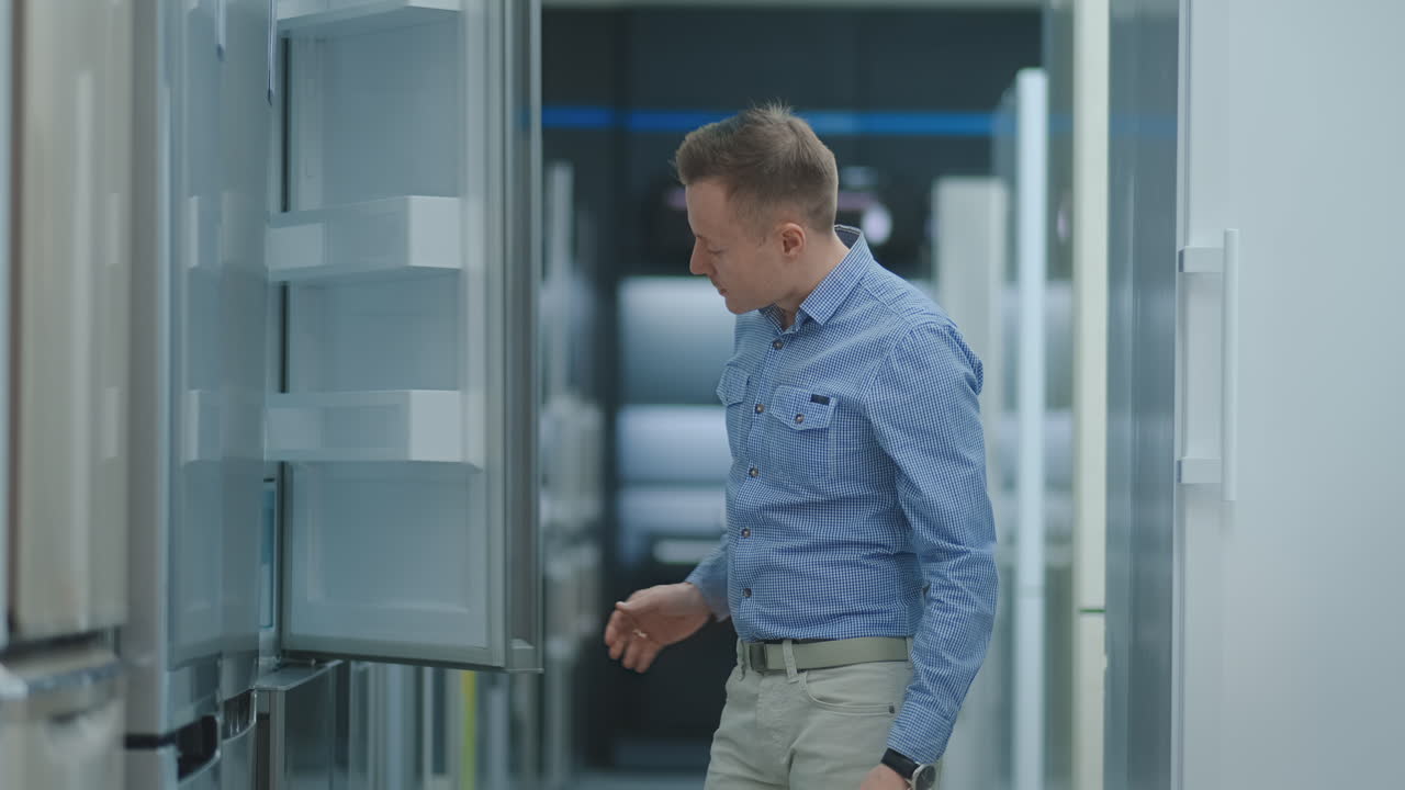 hombre guapo con camisa azul abre la puerta del refrigerador en la tienda de electrodomésticos y comparar con otros modelos para comprar una nueva casa