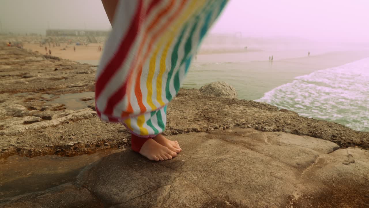 Ung kvinde knæler på et stribet badehåndklæde ved havet.